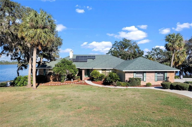 single story home with brick siding, a chimney, a water view, roof mounted solar panels, and a front lawn