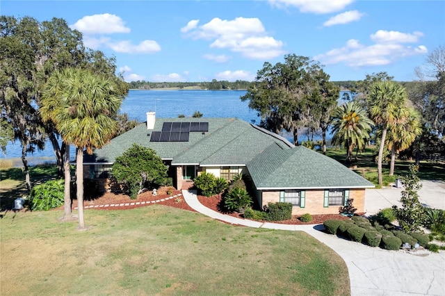 view of front of house featuring roof with shingles, a chimney, solar panels, a water view, and a front yard