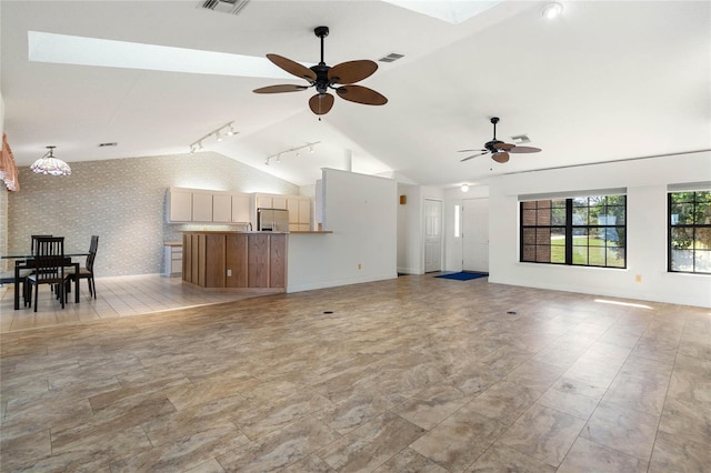 unfurnished living room with ceiling fan, vaulted ceiling with skylight, an accent wall, baseboards, and wallpapered walls