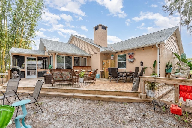 back of property with a sunroom, a chimney, roof with shingles, outdoor dining area, and a wooden deck