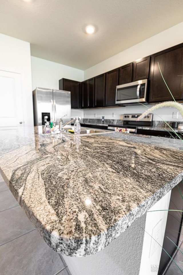 kitchen with stainless steel appliances, dark brown cabinetry, a sink, dark stone counters, and tile patterned floors