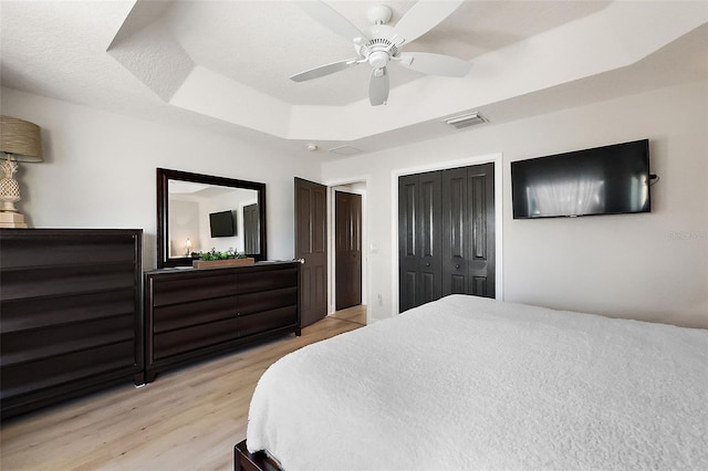 bedroom with visible vents, ceiling fan, a tray ceiling, light wood-style floors, and a closet