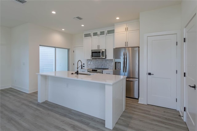 kitchen with stainless steel appliances, backsplash, a kitchen island with sink, white cabinetry, and light wood-type flooring