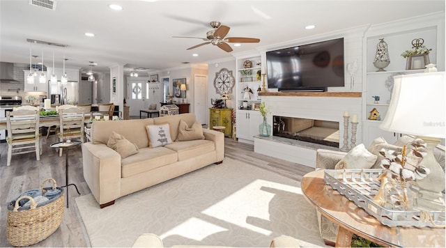 living room with light wood finished floors, a large fireplace, a ceiling fan, crown molding, and recessed lighting