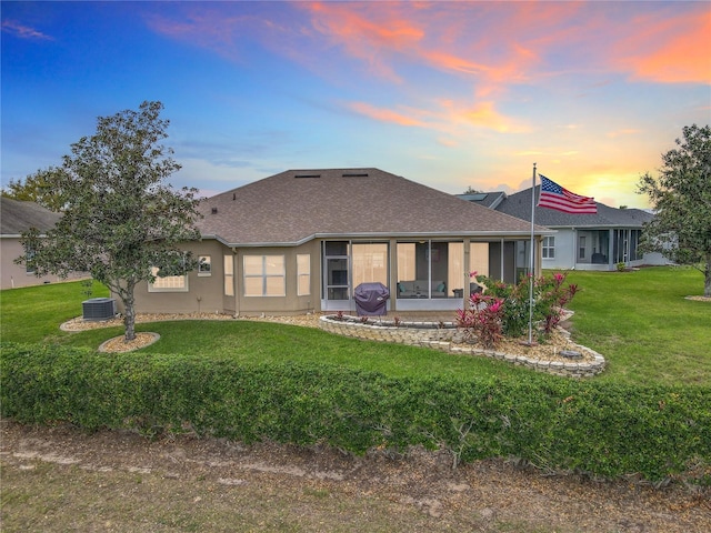 back of property at dusk featuring central air condition unit, a sunroom, roof with shingles, a lawn, and stucco siding