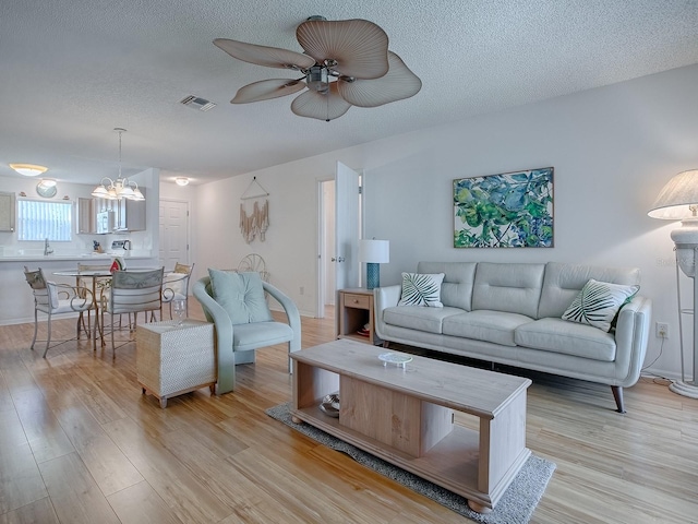 living room featuring visible vents, light wood-style flooring, a textured ceiling, baseboards, and ceiling fan with notable chandelier