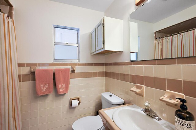 bathroom featuring a shower with shower curtain, toilet, a wainscoted wall, a sink, and tile walls