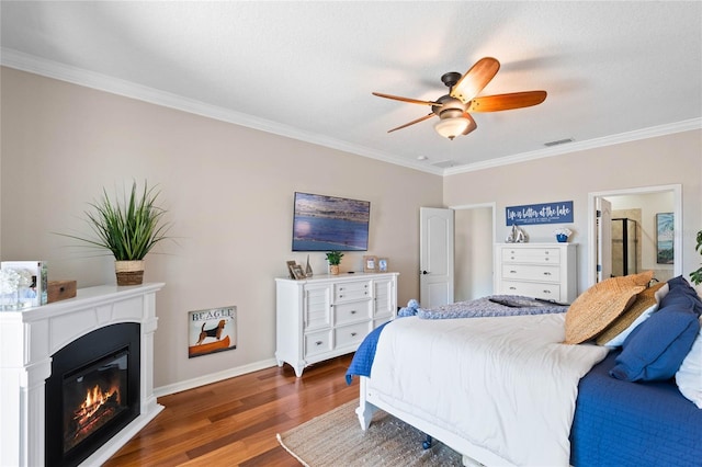 bedroom with wood finished floors, baseboards, visible vents, ornamental molding, and a glass covered fireplace