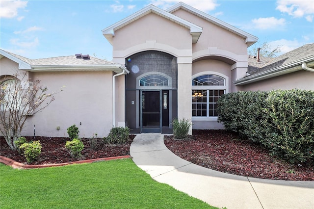 entrance to property with a shingled roof, a lawn, and stucco siding