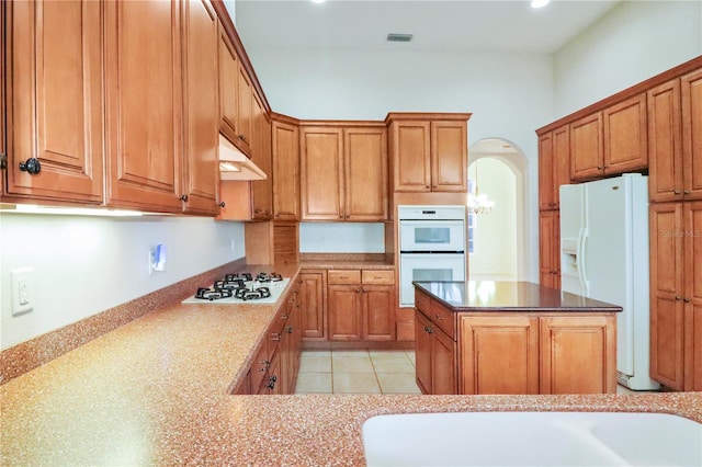 kitchen featuring under cabinet range hood, a center island, white appliances, arched walkways, and brown cabinetry