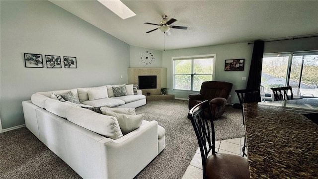 living room featuring a ceiling fan, vaulted ceiling with skylight, a fireplace with raised hearth, a textured ceiling, and light carpet