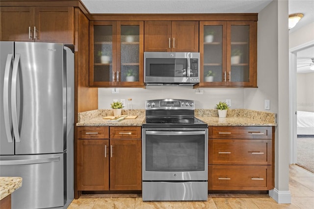 kitchen with light stone counters, stainless steel appliances, glass insert cabinets, and brown cabinetry