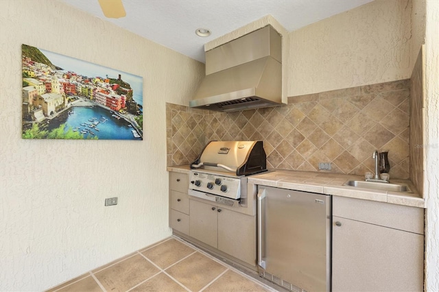 kitchen featuring wall chimney range hood, tile countertops, freestanding refrigerator, light tile patterned flooring, and a sink