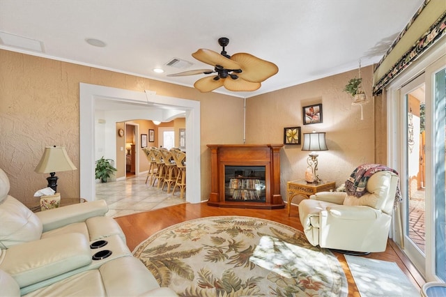 living area with visible vents, ceiling fan, ornamental molding, wood finished floors, and a glass covered fireplace