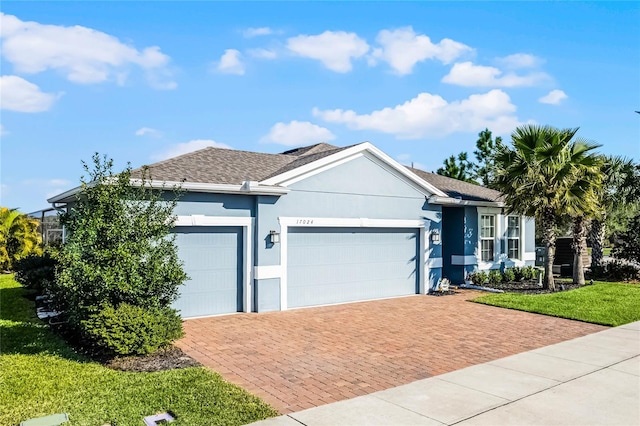 ranch-style house featuring a front yard, an attached garage, a shingled roof, stucco siding, and decorative driveway