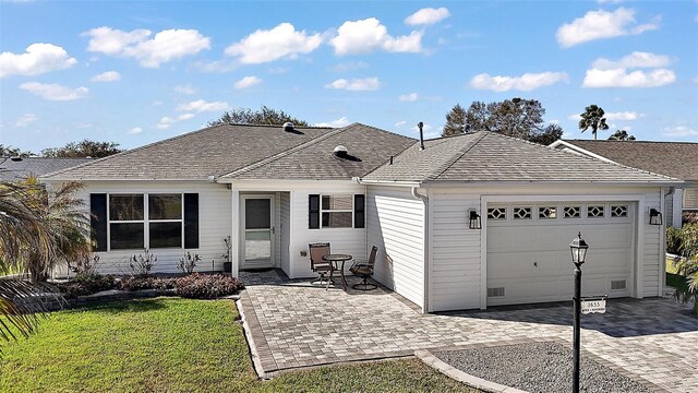 rear view of property featuring decorative driveway, a lawn, a garage, and a shingled roof