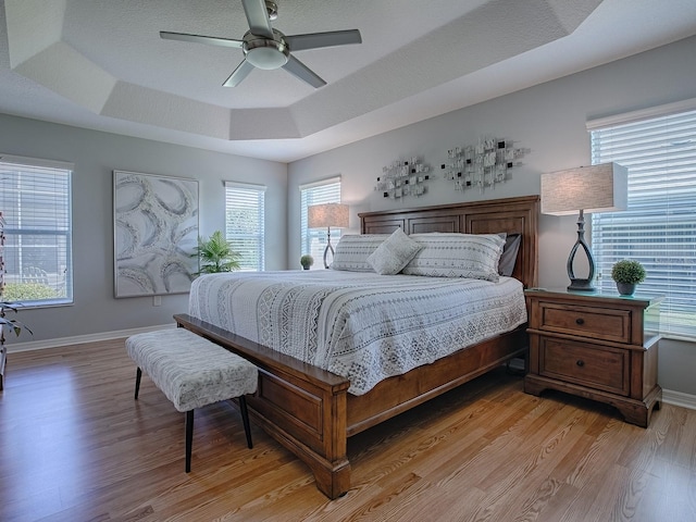 bedroom with multiple windows, a raised ceiling, light wood-style floors, and baseboards