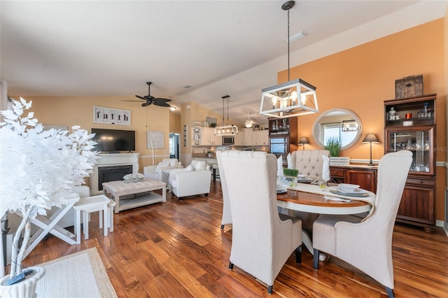 dining area with visible vents, ceiling fan with notable chandelier, a fireplace, dark wood-style flooring, and vaulted ceiling