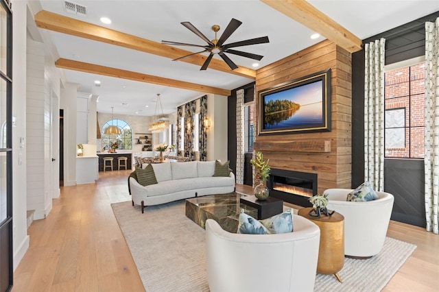 living area with visible vents, light wood finished floors, a glass covered fireplace, beamed ceiling, and ceiling fan with notable chandelier