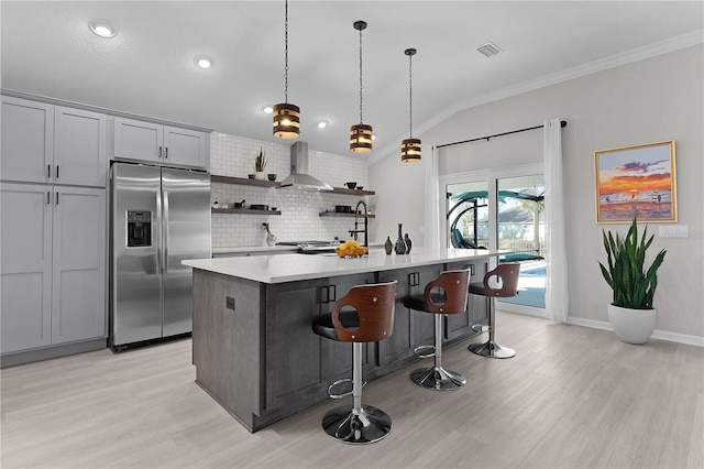 kitchen with open shelves, an island with sink, stainless steel fridge with ice dispenser, light countertops, and wall chimney exhaust hood