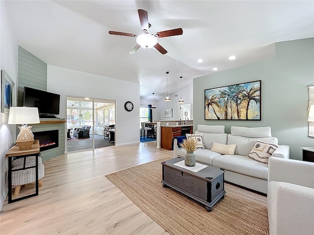 living area with baseboards, lofted ceiling, light wood-style flooring, a warm lit fireplace, and a ceiling fan