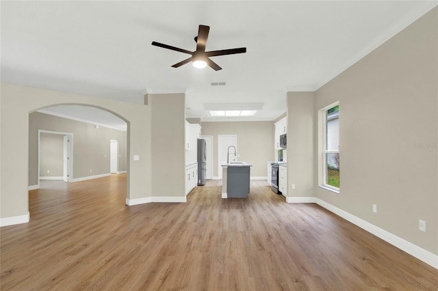 unfurnished living room featuring a ceiling fan, arched walkways, a sink, light wood-style floors, and crown molding