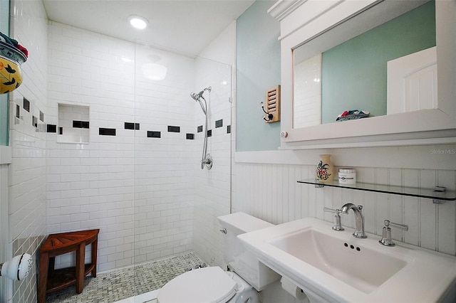 full bathroom with toilet, tiled shower, a wainscoted wall, and a sink