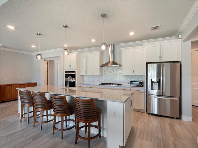 kitchen featuring stainless steel appliances, visible vents, white cabinetry, wall chimney range hood, and an island with sink