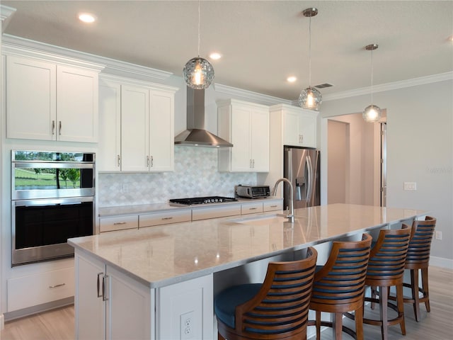 kitchen featuring a kitchen island with sink, a sink, stainless steel appliances, decorative backsplash, and wall chimney range hood