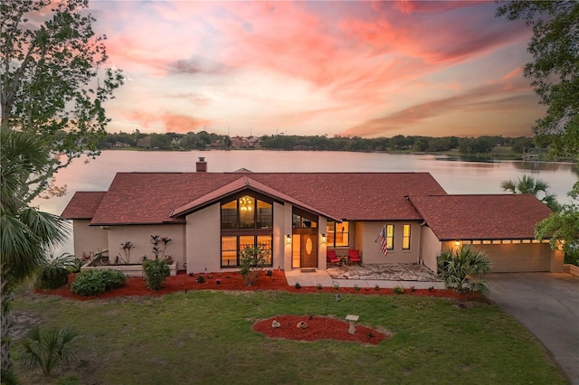 mid-century home featuring a front yard, a chimney, an attached garage, a water view, and driveway
