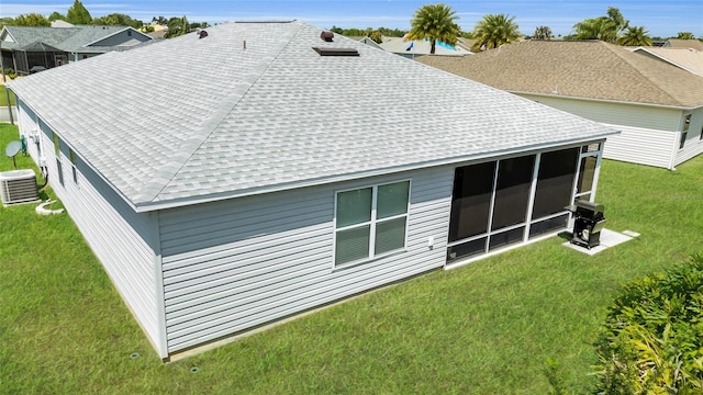 back of property with central AC unit, a shingled roof, a sunroom, and a lawn