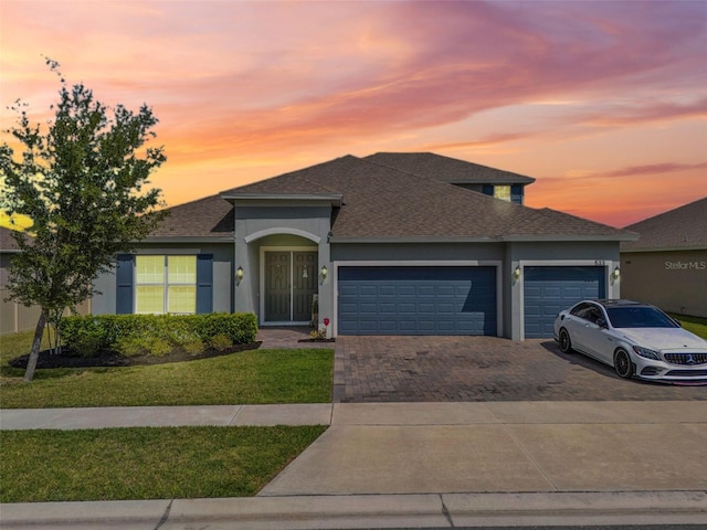 view of front of house with stucco siding, decorative driveway, an attached garage, a front lawn, and a shingled roof