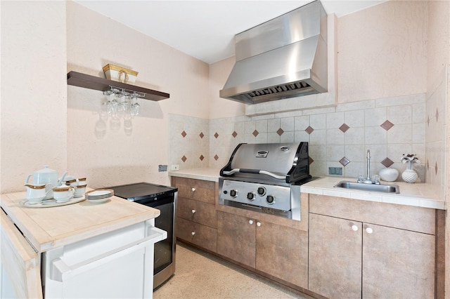 kitchen with wall chimney exhaust hood, open shelves, light countertops, a sink, and decorative backsplash