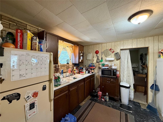 kitchen with a sink, light countertops, freestanding refrigerator, stainless steel microwave, and wood walls