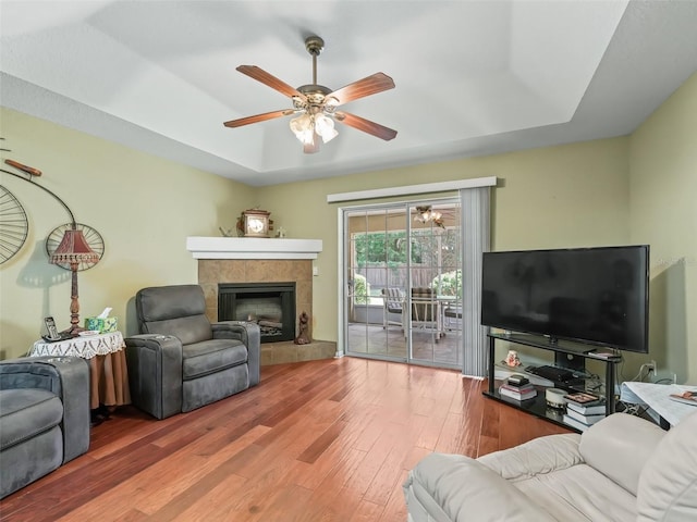 living area with a tile fireplace, ceiling fan, a tray ceiling, and wood finished floors