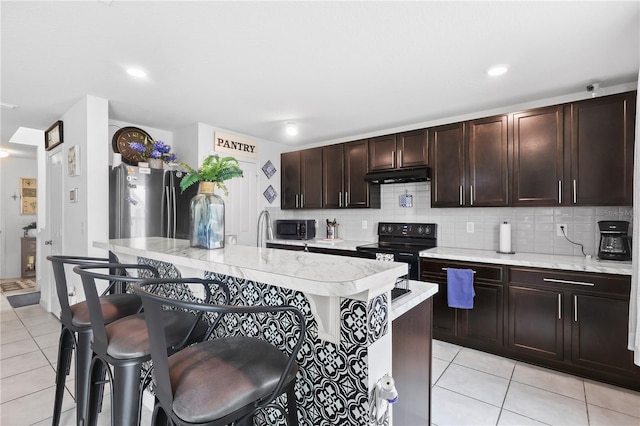 kitchen with decorative backsplash, under cabinet range hood, light tile patterned flooring, a kitchen bar, and electric range