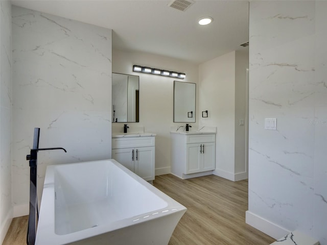 bathroom featuring wood finished floors, a freestanding bath, baseboards, two vanities, and visible vents