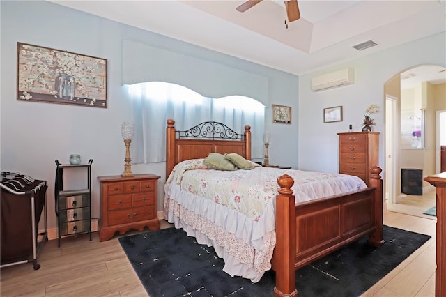 bedroom with light wood-style floors, ceiling fan, arched walkways, visible vents, and a wall unit AC
