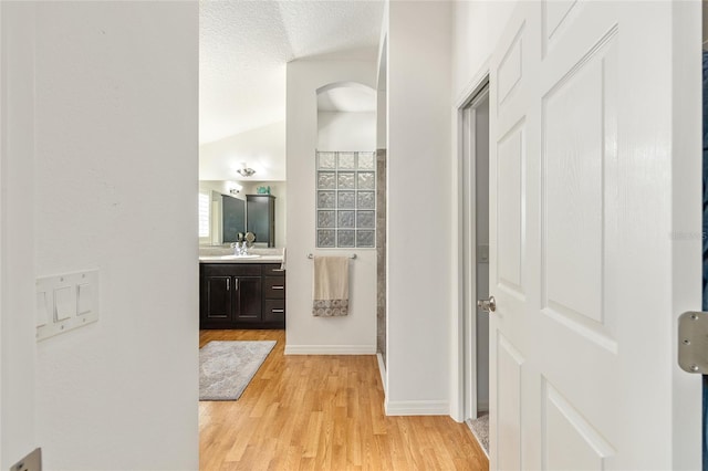 hall featuring a textured ceiling, arched walkways, baseboards, lofted ceiling, and light wood-style floors