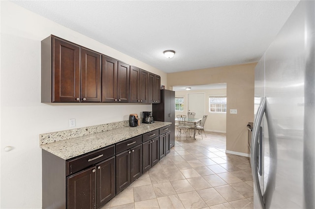 kitchen with light tile patterned floors, freestanding refrigerator, light stone countertops, baseboards, and dark brown cabinetry