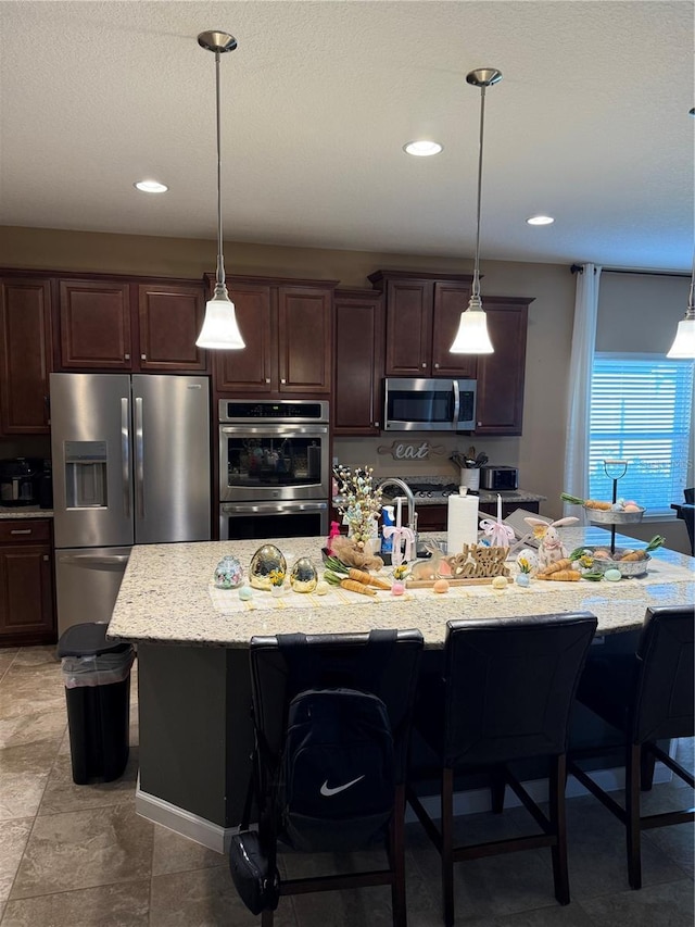 kitchen featuring appliances with stainless steel finishes, a breakfast bar, dark brown cabinetry, a center island with sink, and hanging light fixtures