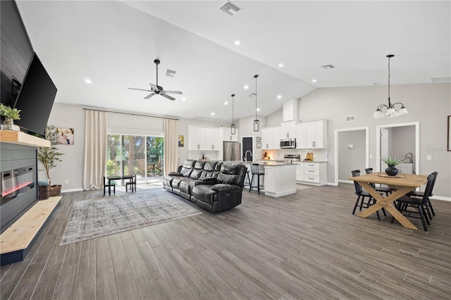 living area featuring visible vents, a glass covered fireplace, wood finished floors, high vaulted ceiling, and ceiling fan with notable chandelier