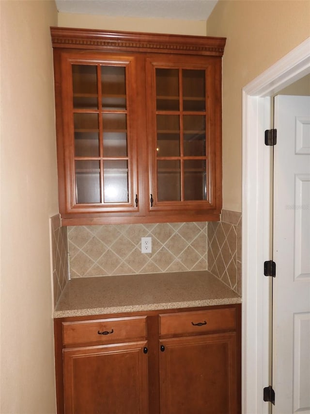 kitchen with glass insert cabinets, light stone counters, decorative backsplash, and brown cabinets