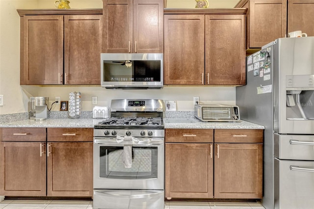 kitchen featuring light tile patterned flooring, light stone counters, brown cabinetry, stainless steel appliances, and a toaster