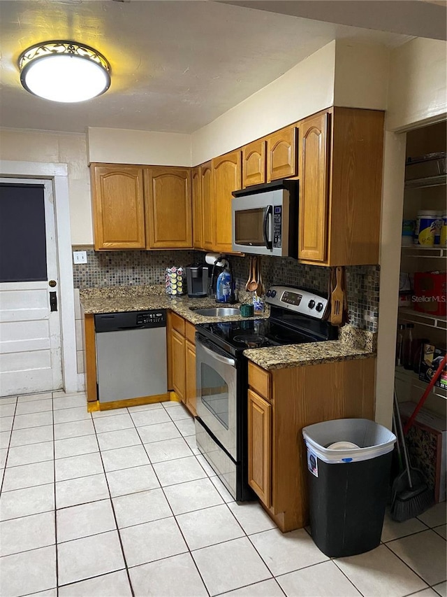 kitchen featuring brown cabinets, light tile patterned flooring, appliances with stainless steel finishes, dark stone counters, and backsplash