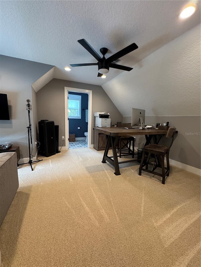 office space featuring a ceiling fan, a textured ceiling, light colored carpet, vaulted ceiling, and baseboards