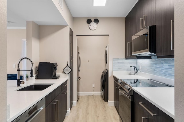 kitchen with a sink, stainless steel appliances, stacked washer / drying machine, light wood-type flooring, and light countertops