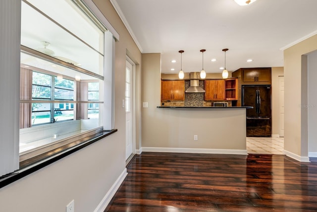 kitchen featuring freestanding refrigerator, wall chimney range hood, brown cabinetry, crown molding, and dark wood-style floors