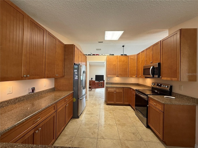 kitchen featuring appliances with stainless steel finishes, brown cabinetry, dark stone counters, a textured ceiling, and light tile patterned flooring