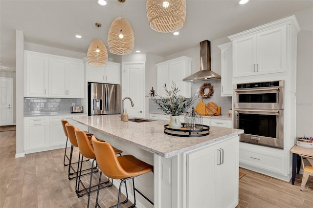 kitchen featuring wall chimney range hood, appliances with stainless steel finishes, a sink, tasteful backsplash, and an island with sink